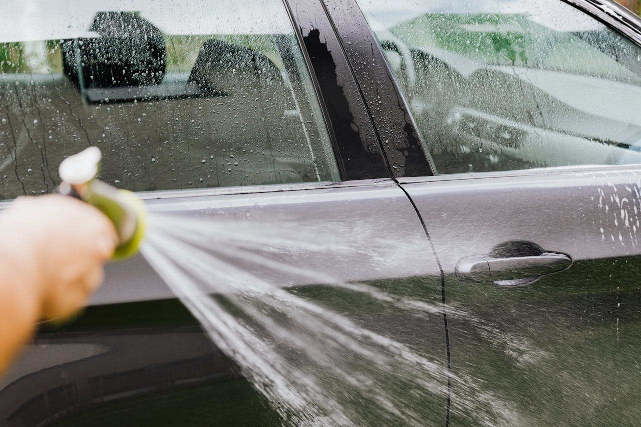 A person uses a hose to wash a black car outdoors, creating water splashes.